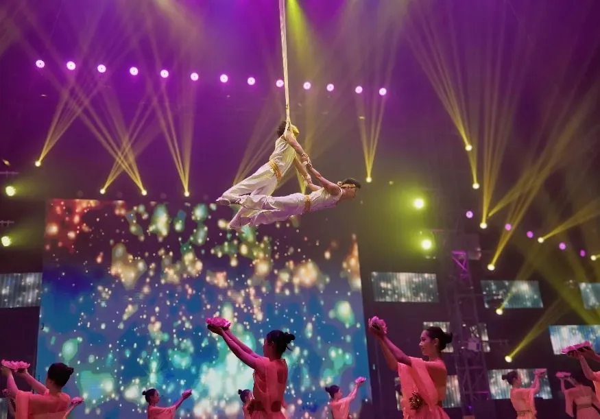 Aerial performers above dancers holding lotus flowers